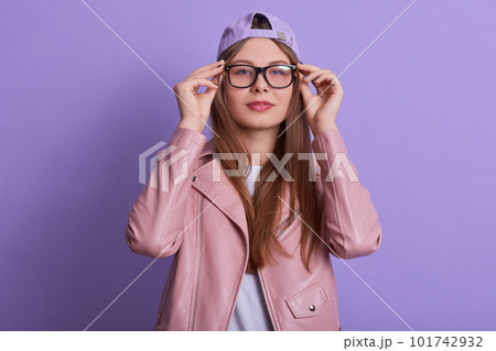 Studio shot of young lady wearing rosy jacket,hat and eyewears, woman keeping hands on frame of glasses, looking at camera, standing with calm facial expression against lilac wall.People concept. Studio shot of young lady wearing rosy jacket,hat and eyewears, woman keeping hands on frame of glasses, looking at camera, standing with calm facial expression against lilac wall.People concept. 101742932