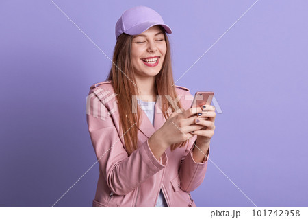 Horizontal shot of pretty blond girl with beautiful long hair, wearing rosy lether jacket and cap, holding smartphone in her hands, cheking social networks. Concept of enjoying life, modern technology Horizontal shot of pretty blond girl with beautiful long hair, wearing rosy lether jacket and cap, holding smartphone in her hands, cheking social networks. Concept of enjoying life, modern technology 101742958