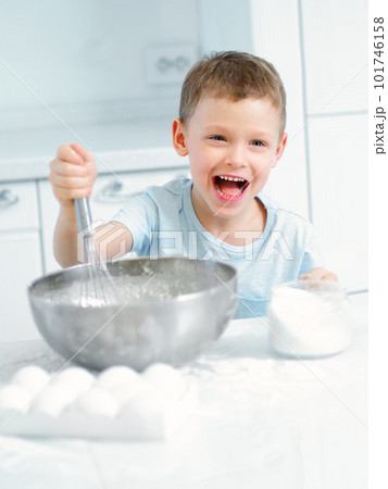 Child cooks food. Caucasian boy sits at table in kitchen and kneads dough in cup with hand whisk Child cooks food. Caucasian boy sits at table in kitchen and kneads dough in cup with hand whisk 101746158