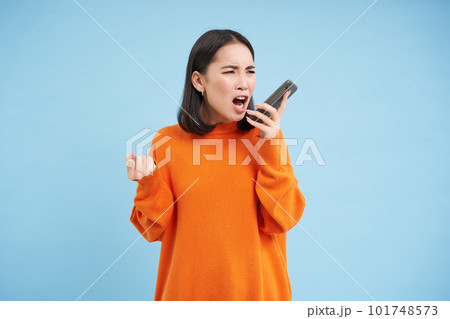Angry asian woman yells at mobile phone, shouts in smartphone with furious face, stands over blue background Angry asian woman yells at mobile phone, shouts in smartphone with furious face, stands over blue background 101748573