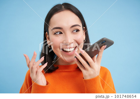 Portrait of asian woman records voice message, talks on speakerphone, holds mobile phone near mouth while speaking, translates her speech on app, blue background Portrait of asian woman records voice message, talks on speakerphone, holds mobile phone near mouth while speaking, translates her speech on app, blue background 101748654