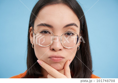 Close up portrait of asian girl in glasses, squints with suspicious face, thinking, stands over blue background 101748708