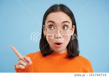 Close up portrait of woman in glasses, points and looks left, shows advertisement, stares at banner with surprised face, blue background Close up portrait of woman in glasses, points and looks left, shows advertisement, stares at banner with surprised face, blue background 101748719