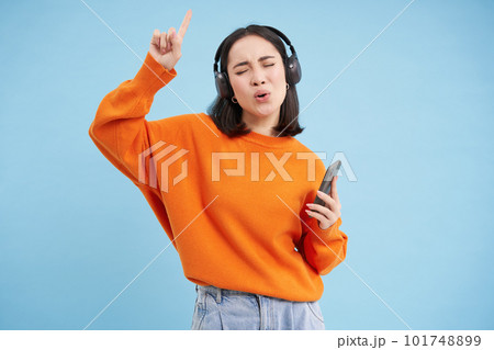 Beautiful korean woman in headphones, dancing, listening music on mobile phone app, smiling, posing over blue background 101748899