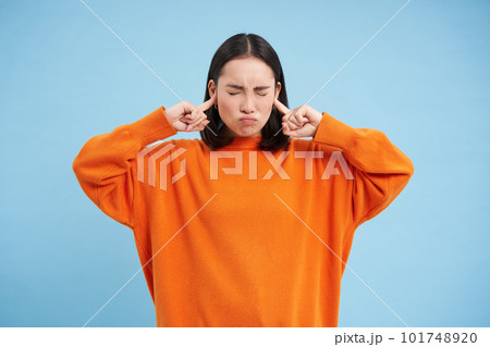 Cant listen. Fed up asian woman shuts her ears with fingers and closes eyes from loud noise, stands in orange sweatshirt over blue background 101748920
