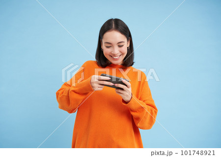 Happy smiling girl, 25 years, plays mobile video games, holds smartphone in both hands, stands against blue background 101748921
