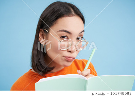 Portrait of asian woman in glasses, making notes in notebook, looks curious and suspicious at camera, stands over blue background Portrait of asian woman in glasses, making notes in notebook, looks curious and suspicious at camera, stands over blue background 101749095