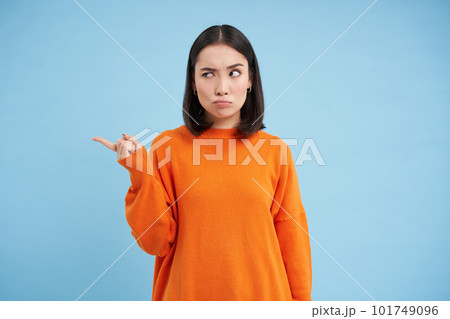 Confused korean girl cant understand something, points left with clueless face, thinking, standing in orange shirt against blue background Confused korean girl cant understand something, points left with clueless face, thinking, standing in orange shirt against blue background 101749096