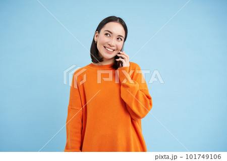Portrait of young candid woman, asian girl talking on cellphone, holds mobile phone and speaks, stands over blue background Portrait of young candid woman, asian girl talking on cellphone, holds mobile phone and speaks, stands over blue background 101749106