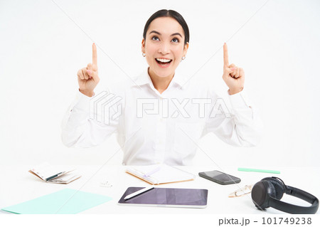 Business and profession. Portrait of asian woman in workplace, sits in office and points up, shows banner advertisement, white background Business and profession. Portrait of asian woman in workplace, sits in office and points up, shows banner advertisement, white background 101749238