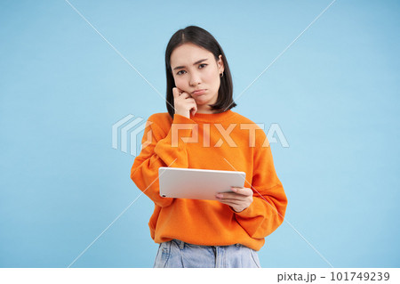 Complicated asian girl, stands with digital tablet with frowning, puzzled face, poses over blue background Complicated asian girl, stands with digital tablet with frowning, puzzled face, poses over blue background 101749239