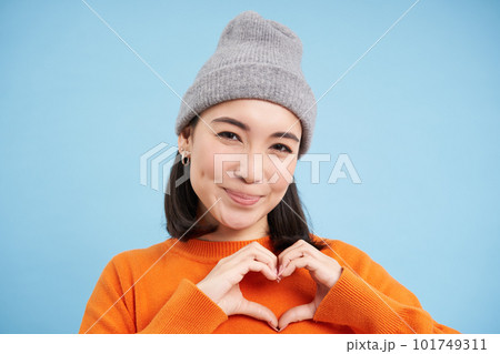 Close up of lovely asian girl in beanie, shows heart sign, looks cute, stands over blue studio background 101749311