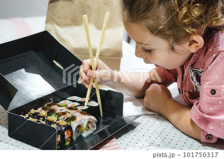 A cute white Caucasian little girl picking up some sushi maki for lunch with chopsticks from a takeaway box on the kitchen table at home 101750317