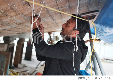 A middle-aged or young man working on the hull of a boat - putting cotton caulk into the cracks between wooden planks (caulking) 101750321