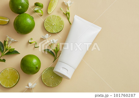 Against a beige background, some Lime (Citrus aurantiifolia) slices, peels and flower branches are arranged with a white tube. Product mockup with empty label. copy space in the right side 101760287