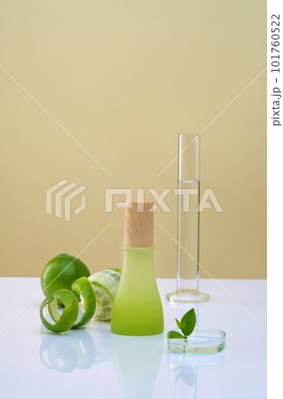 Front view of green bottle with wooden cap unlabeled, lab glassware, fresh limes and spiral peel decorated on color background. Mockup for product with ingredients from lemon essential oil. 101760522