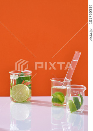 Lime slices, peel and green leaves in beakers and colorless liquid decorated on orange neon background. Scene for advertising cosmetic of natural extract. Blank space for display Lime slices, peel and green leaves in beakers and colorless liquid decorated on orange neon background. Scene for advertising cosmetic of natural extract. Blank space for display 101760536