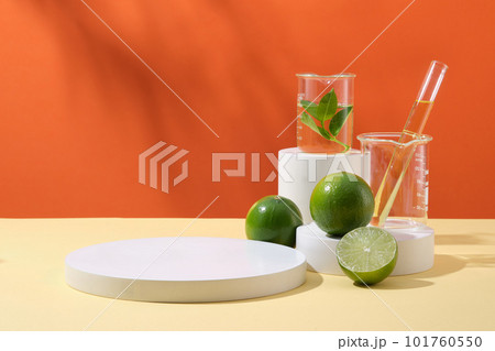 Pedestal for cosmetic product display presentation with round empty podium, limes and lab glassware containing yellow liquid on orange background. Advertising photo, copy space 101760550