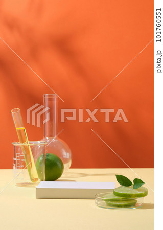 Front view of lime slices on petri dish, lab glassware containing yellow liquid and white podium on orange background. Empty space for cosmetic of lime extract presentation. 101760551