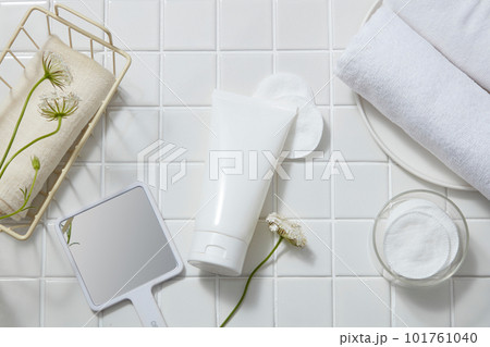 Over white tile background, an unlabeled tube is decorated with cotton pads, flower branches, mirror, towels. Product mockup of Aloe vera extract. It helps to moisturize the skin 101761040