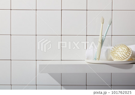 Bathroom background with toothbrushes in glass cup, scalp massage brush and towel on white pedestal on tile wall background. Empty space for display cosmetic product. Front view, copy space. 101762025