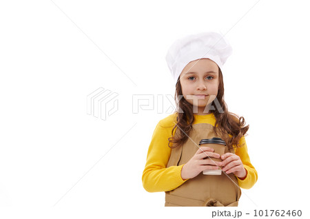 Lovely child girl in chef's hat and apron. looking at camera, holding disposable recyclable paper cup with takeaway drink, isolated on white background. Copy space. Food and drink delivery service 101762460