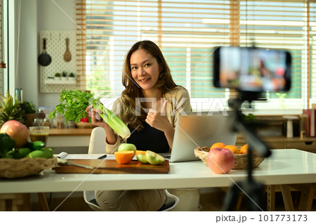 Confident female nutritionist holding celery, providing with online consultations or recording video content on smartphone 101773173