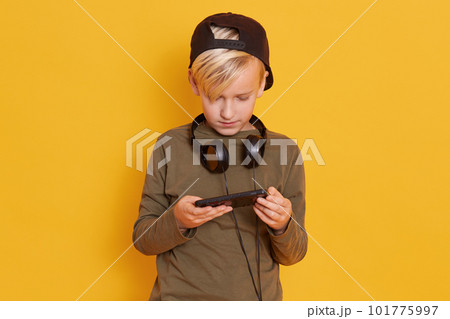 Portrait of little blond kid playing video game and holding smartphone in hands, keeps heandphones around neck, dresses green shirt and black cap, model posing isolated over yellow background. 101775997