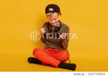 Photo of delighted little boy dresses orange trousers and green shirt playing video game on cellphone and clenching fist while sitting on floor with crossed legs isolated over yellow studio background 101776002
