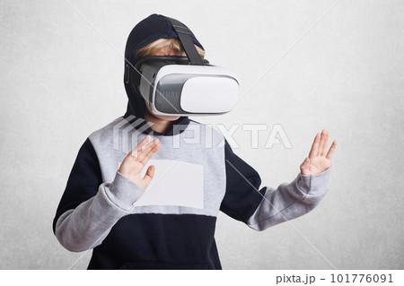 Photo of small kid wears virtual relality goggles, experiences something excited, gestures, entertains himself. Child in VR glasses with arms stretched forward and palms up, gropes something invisable 101776091