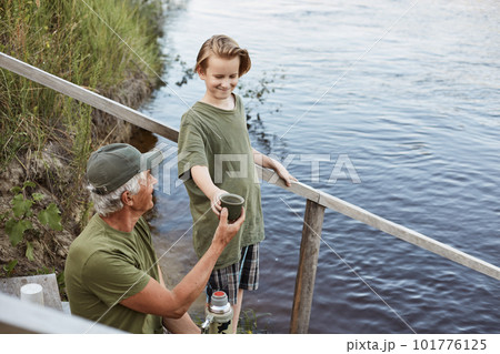 Happy family drinking tea from thermos while enjoying fishing, grandfather and grandson posing on wooden stairs leading to water, father and son wearing green t shirts. Happy family drinking tea from thermos while enjoying fishing, grandfather and grandson posing on wooden stairs leading to water, father and son wearing green t shirts. 101776125