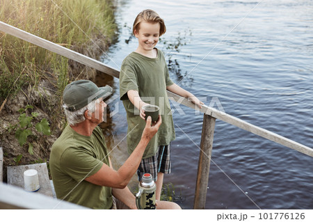 Father and son spending time together at bank of river or lake, senior man giving cup of tea from thermos to his grandson, family posing on wooden stairs leading to water, rest on beautiful nature. Father and son spending time together at bank of river or lake, senior man giving cup of tea from thermos to his grandson, family posing on wooden stairs leading to water, rest on beautiful nature. 101776126
