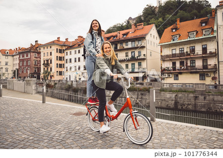 Couple of best friends riding a bike on the city streets, one of the women standing on the trunk of the bike  101776344