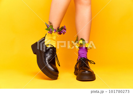 Close up portrait of tender female feet wearing black boots and multi colored socks with flowers inside over yellow studio background. Wildflowers 101780273