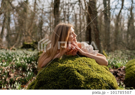 Snowdrops galanthus blond. A girl in a white dress lay down on a stone in the moss in a meadow with snowdrops in a spring forest Snowdrops galanthus blond. A girl in a white dress lay down on a stone in the moss in a meadow with snowdrops in a spring forest 101785507