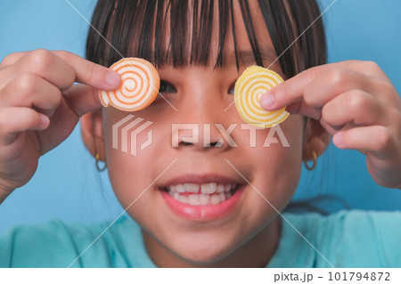 Smiling cute little girl covering her eyes with two gelatinous sweets isolated on blue background. Children eat sugary sweets, causing loss teeth or tooth decay and unhealthy oral care. Smiling cute little girl covering her eyes with two gelatinous sweets isolated on blue background. Children eat sugary sweets, causing loss teeth or tooth decay and unhealthy oral care. 101794872
