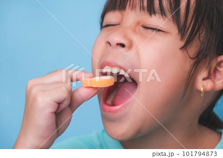 Smiling cute little girl eating sweet gelatin with sugar added isolated on blue background. Children eat sugary sweets, causing loss teeth or tooth decay and unhealthy oral care. Smiling cute little girl eating sweet gelatin with sugar added isolated on blue background. Children eat sugary sweets, causing loss teeth or tooth decay and unhealthy oral care. 101794873