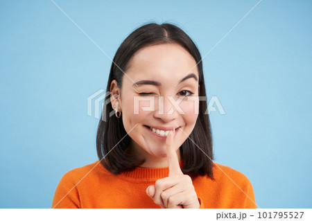 Close up of smiling asian girl, winks at you, tells secret, holds finger on lips, stands over blue studio background Close up of smiling asian girl, winks at you, tells secret, holds finger on lips, stands over blue studio background 101795527
