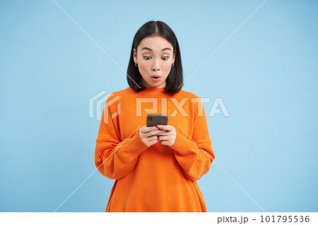 Young woman looks amazed at her cellphone, reads exciting news, found good deal online on mobile application, standing over blue background 101795536