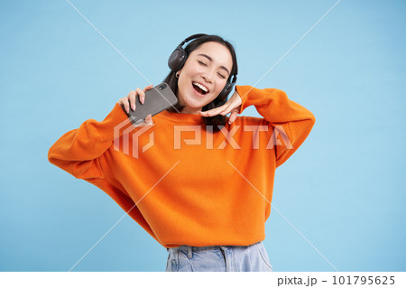 Beautiful happy korean woman in headphones, listening music on mobile phone in earphones, wearing orange sweatshirt, blue background Beautiful happy korean woman in headphones, listening music on mobile phone in earphones, wearing orange sweatshirt, blue background 101795625