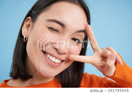 Close up portrait of positive asian woman, winks and shows peave, v-sign gesture, sticks tongue and smiles, stands over blue background 101795938