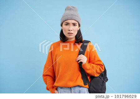 Young asian woman in hat, holds backpack, sulks and looks confused, perplexed emotion, stands over blue background 101796029