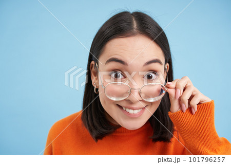 Portrait of brunette japanese woman in glasses, smiling and looking with happy face at camera, tries prescribed eyewear, blue background Portrait of brunette japanese woman in glasses, smiling and looking with happy face at camera, tries prescribed eyewear, blue background 101796257