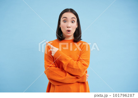 Surprised asian girl points left, shows advertisement with amazed face, shocked by promo offer aside on blue background 101796287