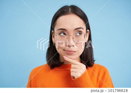Portrait of thoughtful korean woman in glasses, thinking and looking aside, has an idea, plans smth, stands over blue background 101796628