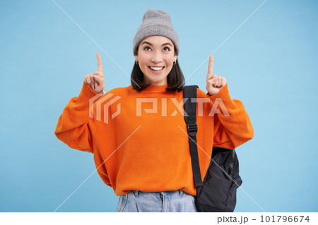 Smiling beautiful korean woman, pointing finger up, shows advertisement on top, wears street outfit and backpack, blue background 101796674
