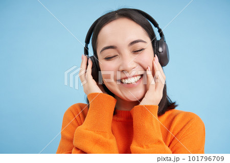 Close up of beautiful asian woman in headphones, smiles and listens music, enjoys podcast, stands over blue background Close up of beautiful asian woman in headphones, smiles and listens music, enjoys podcast, stands over blue background 101796709