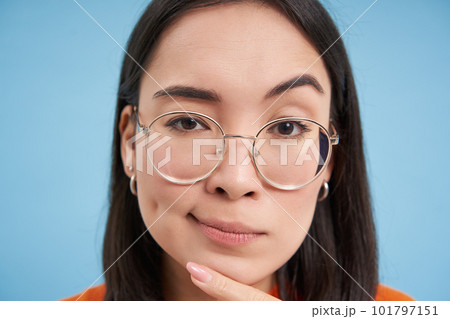Close up portrait of asian woman looks intrigued, wears glasses, squints thoughtful, thinking, making assumption, standing over blue background Close up portrait of asian woman looks intrigued, wears glasses, squints thoughtful, thinking, making assumption, standing over blue background 101797151