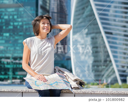 Smiling woman is reading paper map sitting on background of buildings with glass walls. Travel around city. Urban tourism. Modern architectural landmarks. 101800982