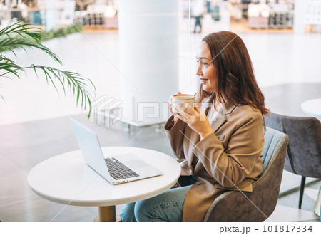 Adult woman with long hair in stylish beige suit working on laptop drinking coffee at cafe 101817334
